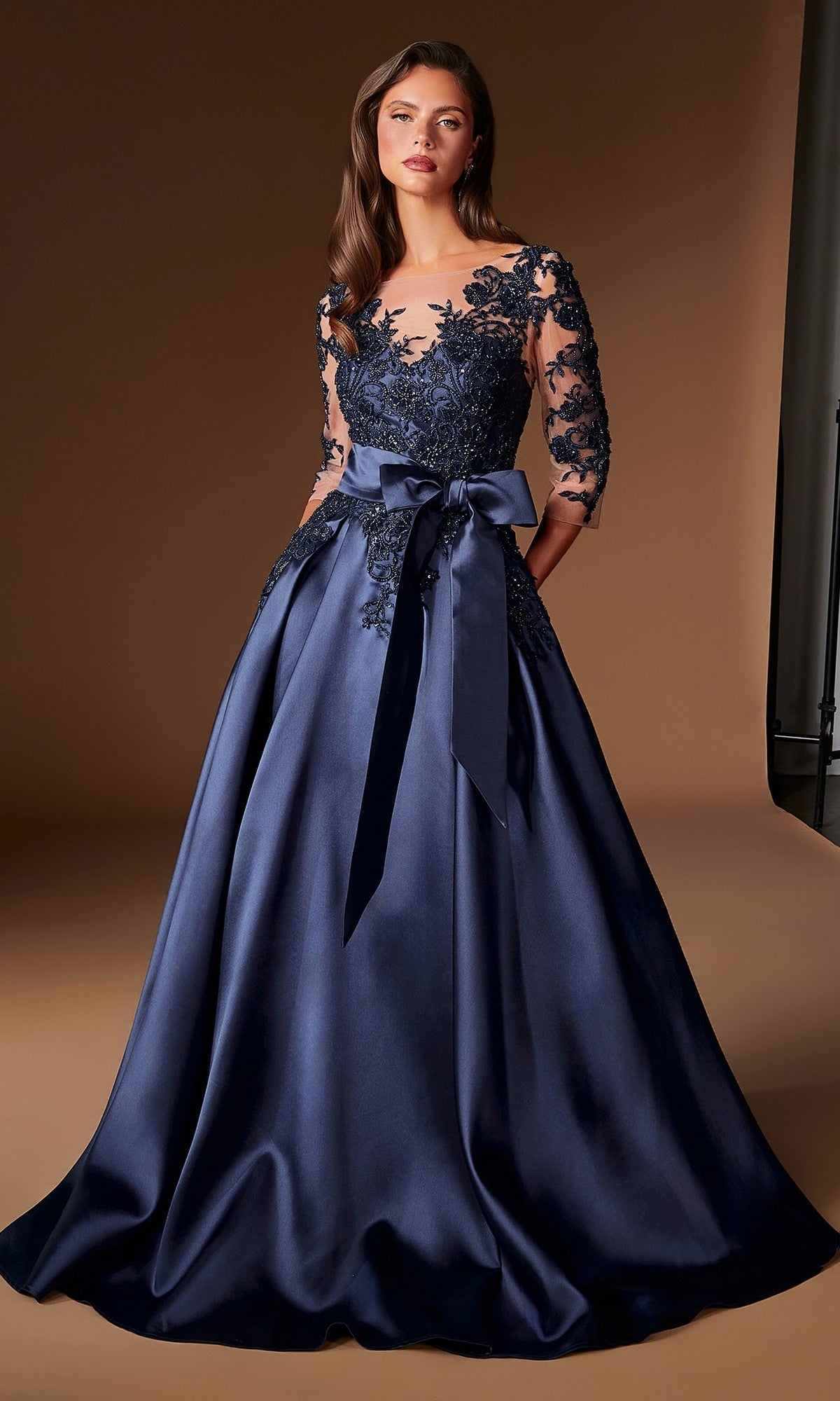 A woman is wearing the Ladivine CDS569 Long Formal Dress, a floor-length navy blue evening gown featuring embroidered lace detailing on the bodice and sleeves, and a large satin bow at the waist, as she stands against a plain brown background.